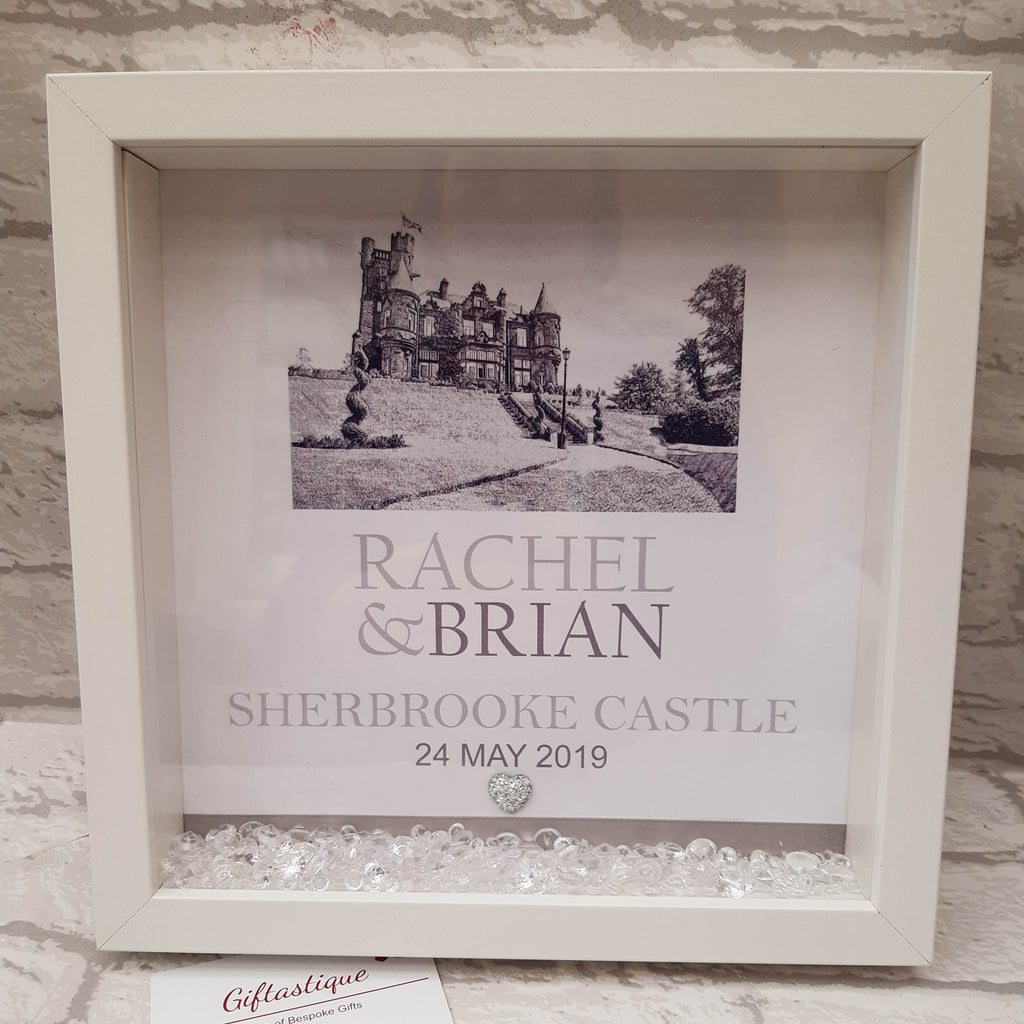 A white square shadow box frame displayed against a white brick wall, containing a black-and-white castle photo, the names 'Rachel & Brian,' 'Sherbrooke Castle,' and the date '24 May 2019,' with small clear stones at the bottom.