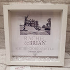 A white square shadow box frame displayed against a white brick wall, containing a black-and-white castle photo, the names 'Rachel & Brian,' 'Sherbrooke Castle,' and the date '24 May 2019,' with small clear stones at the bottom.