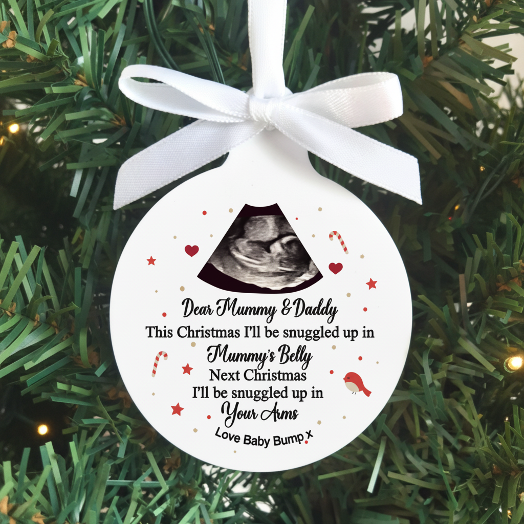 A round white Christmas ornament with a white ribbon bow, featuring a black ultrasound image and a personalized message for 'Mummy & Daddy' about being snuggled up in 'Mummy's Belly' and 'Your Arms'. The ornament is displayed hanging against a green pine tree background.