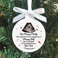 A round white Christmas ornament with a white ribbon bow, featuring a black ultrasound image and a personalized message for 'Mummy & Daddy' about being snuggled up in 'Mummy's Belly' and 'Your Arms'. The ornament is displayed hanging against a green pine tree background.