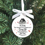 A round white personalized Christmas ornament hanging from a white ribbon bow against a green pine tree background. The ornament features a black ultrasound photo, red stars and hearts, and a sentimental quote for 'Daddy' about being snuggly in 'Mummy's Belly' and 'Your Arms' with 'Love Baby Bump'.