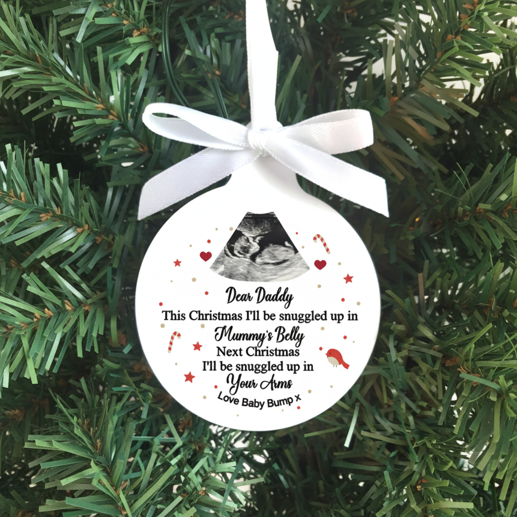 A round white personalized Christmas ornament hanging from a white ribbon bow against a green pine tree background. The ornament features a black ultrasound photo, red stars and hearts, and a sentimental quote for 'Daddy' about being snuggly in 'Mummy's Belly' and 'Your Arms' with 'Love Baby Bump'.