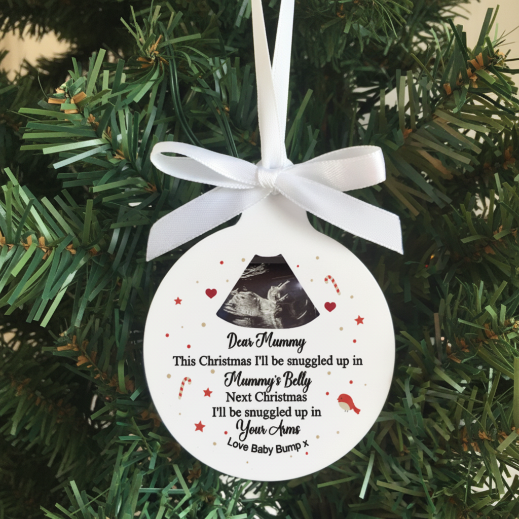 A round white personalized baby bump Christmas ornament hanging on a green Christmas tree. The ornament features a white ribbon bow at the top, a black photo window showing an ultrasound, and printed black text with red and gold accents.